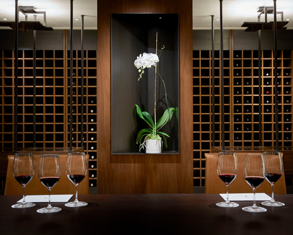 The library at Opus One with three bottles of Opus One and a tasting set up for two guests, including three glasses of wine each. There is a white orchid and a cellar of wine bottles in the background