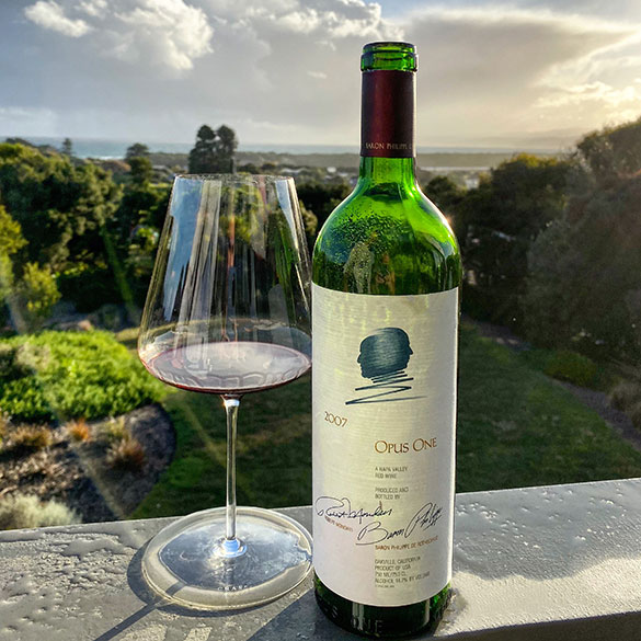 Bottle of Opus One 2007 next to a red wine glass, with clouds and sunshine in the background