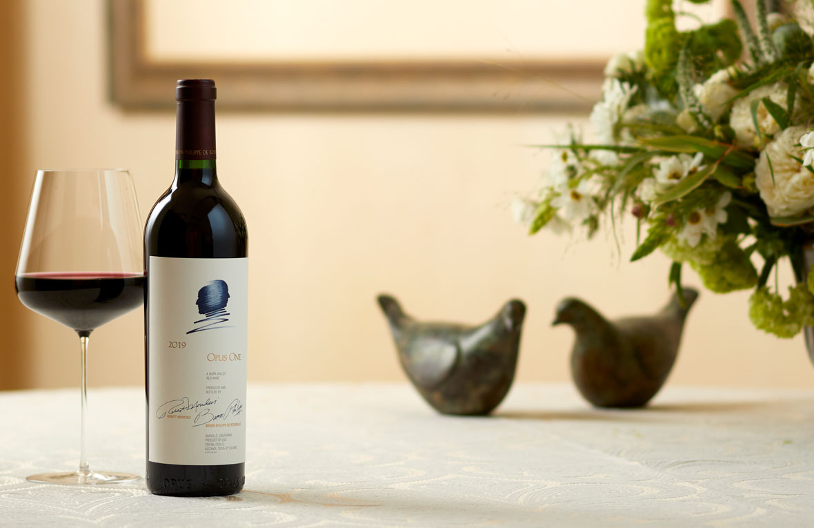 A bottle of Opus One 2019 with two dove statues and green and white florals in the background