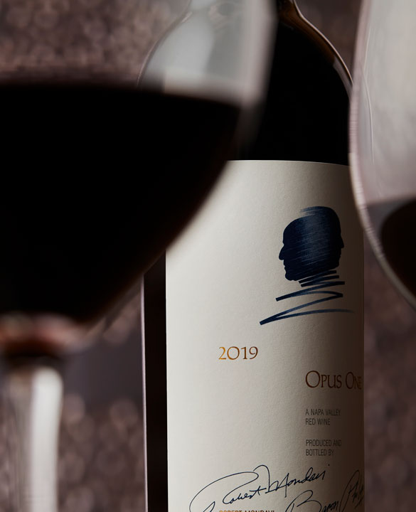 A bottle of Opus One 2019 with two glasses of red wine, and an artistic background